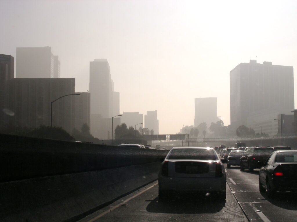 A perspective from a car on a highway, showing traffic and a hazy city skyline. Smog obscures much of the view of city. 