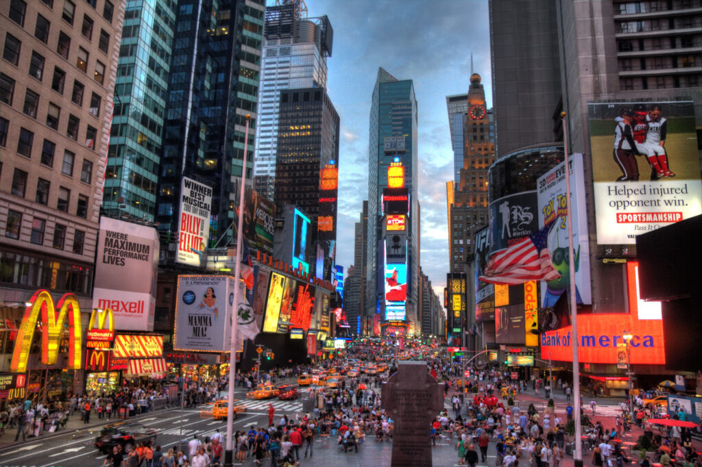 A view of Times Square in New York City from just above ground level. Buildings rise up above the picture taker, and many people walk on the street below. The overall lighting is dim, with bright storefront signs and billboards scattered throughout. 