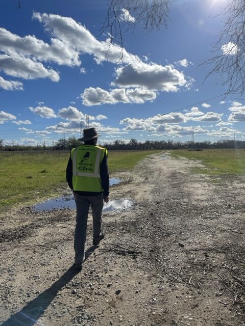 Jacob, wearing a yellow vest, walks along a dirt path with grassy fields to either side and the city skyline faintly visible in the distance. 