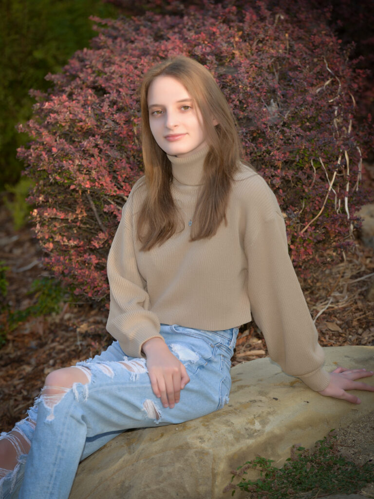 Haley sits on a rock, with a red-leaved bush in the background. She wears a tan sweater and blue jeans. 