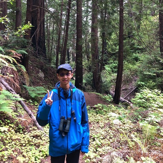 Jih-Heng stands in a redwood forest, giving a peace sign to the camera, with a pair of binoculars hanging around his neck. He wears a ball cap and a bright blue rain jacket.