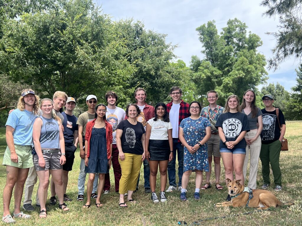 A group of people pose, smiling, in a grassy field. The three team leads stand to the far left, Professor Patricelli stands in the front middle, and the undergraduate team members form the rest of the group. Team mascot Poppy (brown dog) lays in the front right.