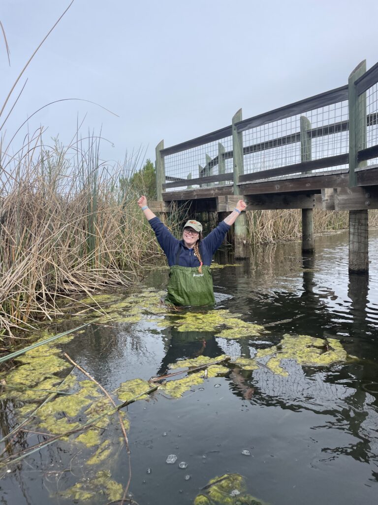 A person stands in a pond with waist high water, wearing green waders, raising her arms above her head, and smiling at the camera.